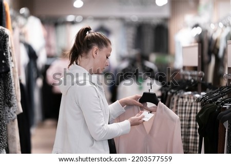 A young pretty woman in white clothes buys clothes in a boutique store, looks at the prices on the label , lifestyle real life photo clothing selection new collection
