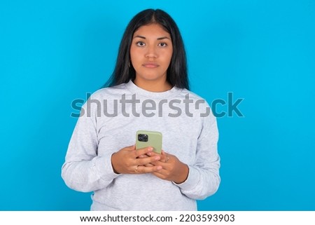 Portrait of serious confident young latin woman wearing grey sweatshirt over blue background holding phone in two hands