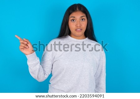 Emotive young latin woman wearing grey sweater over blue background keeps jaw dropped from shock demonstrates amazing promo points right on blank space demonstrates big shopping sale. Advertisement