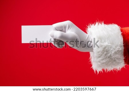 Image of hand of santa claus holding white card with copy space on red background. Christmas, tradition and celebration concept.