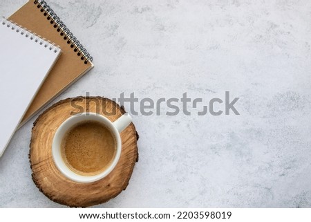 Cup of cappuccino coffee on tree cut wooden cup coaster, spiral notebook and kraft paper sketchbook on office table background. Top view, flat lay, copy space. Stationery supply 