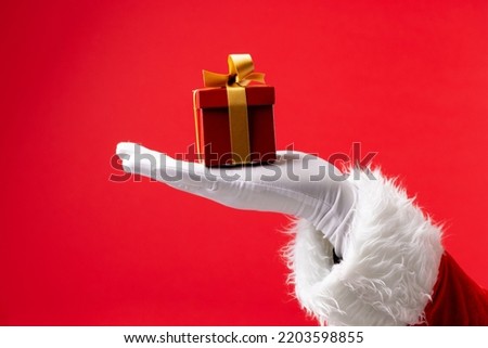 Image of hand of santa claus holding christmas gift with copy space on red background. Christmas, tradition and celebration concept.