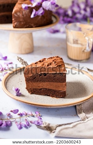 Slice of cake standing on a plate, chocolate brownie cake, with purple flowers around, and a cup of coffee