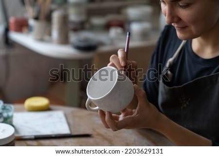 Happy craftswoman wants to decorate kitchen with handmade clay dish with patterns. Lady master enjoys making dish using pencil to draw sketch in studio. Young woman wearing apron draws picture