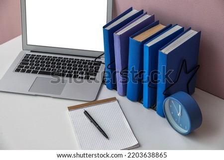 Holder with books, alarm clock and laptop on table near pink wall