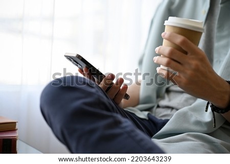 A casual young Asian man using his smartphone and sipping coffee while relaxing in the cafe. close-up image