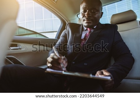 Business concept of a handsome dark-skinned man in a suit and glasses in the back seat of a luxury car signing documents and making work notes