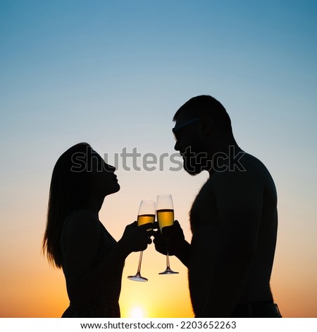 silhouettes of man and woman at sunset dramatic sky background, couple toasting wine glasses in romantic date setting, looking each other, smiling and holding in their hands glasses of champagne.