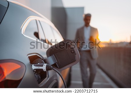 Close up of electric car charging, businessman standing in background and waiting. Royalty-Free Stock Photo #2203712715