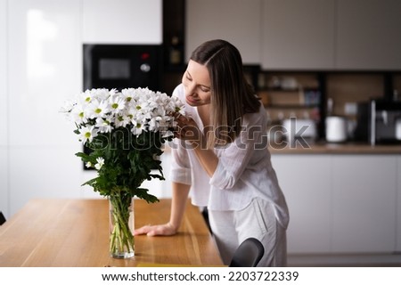 Happy and joyful young woman in white arranging white flowers at home in the kitchen Royalty-Free Stock Photo #2203722339