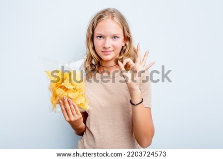 Caucasian teen girl holding bag of chips isolated on blue background cheerful and confident showing ok gesture.