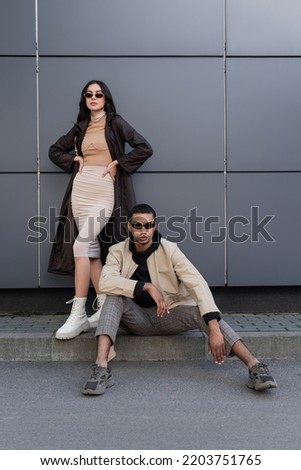 full length of stylish multiethnic couple in trendy autumnal outfits and sunglasses posing near building Royalty-Free Stock Photo #2203751765