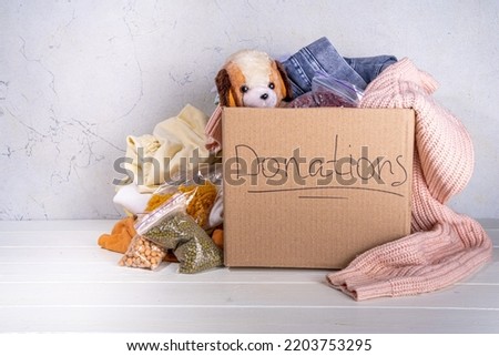 Donation box with clothes, toys and food on white table background. Cardboard donation box full with toys, female and child clothes, foods. Social care, volunteering, charity concept copy space