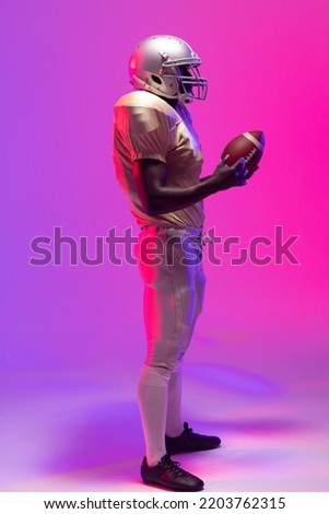 African american male american football player holding ball with neon purple and pink lighting. Sport, movement, training and active lifestyle concept.