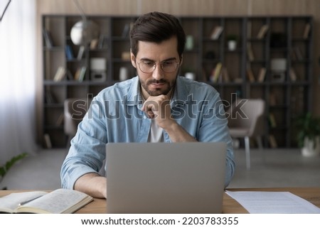 Learning subject online. Serious millennial bearded male in glasses watch elearning training course webinar at laptop screen from home. Pensive young man get remote education study video lesson on pc Royalty-Free Stock Photo #2203783355