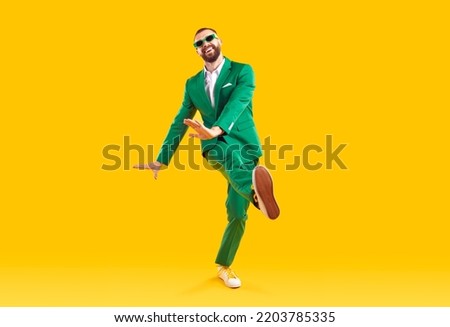 Funny young guy in a party suit dancing in a modern studio. Full body shot of a happy, cheerful, goofy man wearing a green suit and sunglasses dancing isolated on a bright yellow colour background