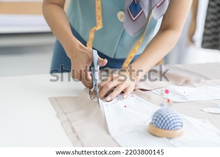 Female dressmaker using scissors cutting fabric dress on the sketch lines in the sewing workshop. Hands of dressmaker cutting dress detail on the sketch lines in tailor shop Royalty-Free Stock Photo #2203800145