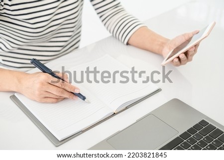 Hands writing notes in notepad while working on the phone and laptop , close up clean image in front of the window in soft light