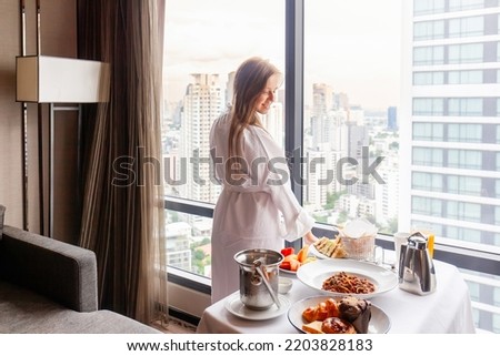 Room service in luxury hotel. Woman in bathrobe eating breakfast near window with city megapolis view. Served food on table in modern cozy room in hotel. Vacation or holidays concept Royalty-Free Stock Photo #2203828183