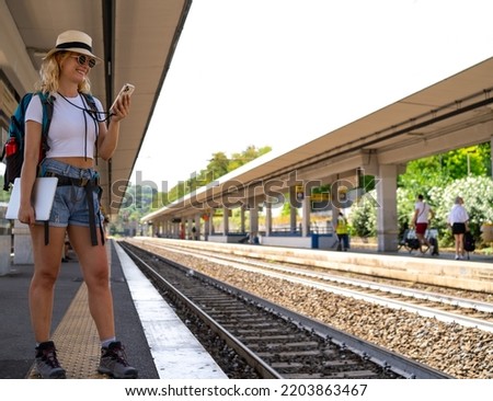 Woman waits for train at station while working happily on phone, digital nomad, travel and transportation concept