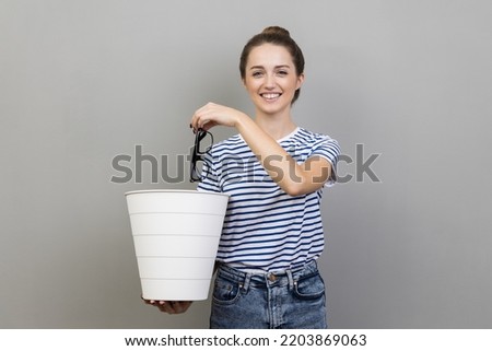 Portrait of smiling positive pretty woman wearing striped T-shirt throwing out eyeglasses to white bin, vision treatment, laser correction. Indoor studio shot isolated on gray background. Royalty-Free Stock Photo #2203869063