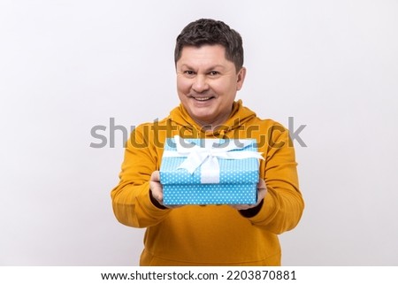 Portrait of positive generous man smiling and giving gift box, sharing holiday present, charity concept, wearing urban style hoodie. Indoor studio shot isolated on white background.