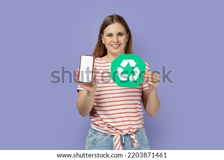 Portrait of cheerful pretty blond woman wearing striped T-shirt standing holding recycling sign and smart phone with blank screen for advertisement. Indoor studio shot isolated on purple background.