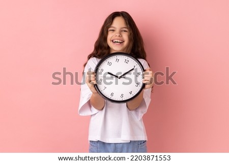 Portrait of smiling delighted little girl wearing white T-shirt holding wall clock, being happy, deadline, satisfied with completed home task. Indoor studio shot isolated on pink background.