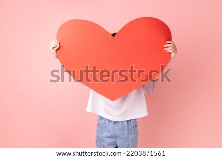 Portrait of anonymous little girl wearing white T-shirt hiding his face behind big red paper heart, making anonymous surprise, sending greeting card. Indoor studio shot isolated on pink background.