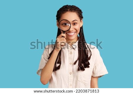 Portrait of funny woman with black dreadlocks holding magnifying glass and looking at camera with big zoom eye, positive face, wearing white shirt. Indoor studio shot isolated on blue background.