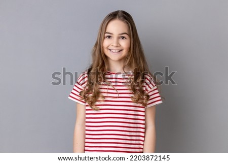 Portrait of satisfied joyful little girl wearing striped T-shirt standing and looking smiling at camera, expressing positive emotions. Indoor studio shot isolated on gray background. Royalty-Free Stock Photo #2203872145
