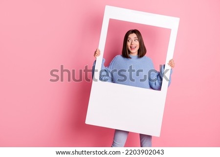 Photo of young funny beaming girl holding white photo frame want more likes looking empty space isolated on pink color background