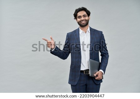 Smiling young indian business man, arab professional manager, eastern businessman executive wearing suit standing holding laptop pointing advertising isolated on beige background, portrait. Royalty-Free Stock Photo #2203904447