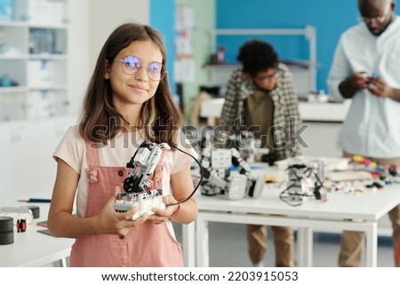 Happy schoolgirl in eyeglasses and casualwear holding handmade robot and looking at camera against her teacher and classmate Royalty-Free Stock Photo #2203915053