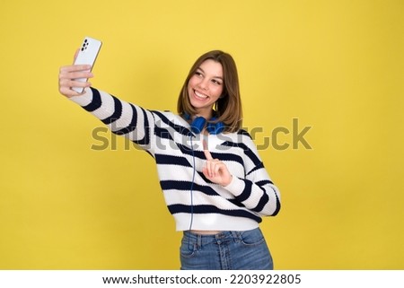 Funny teenage  girl making selfie. Smiling girl wearing  holding  smartphone, making faces on camera, posing for selfie . yellow  background