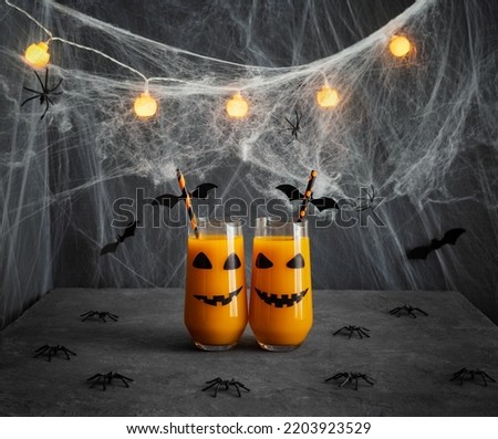 Halloween pumpkin or carrot drink in glasses with jack-o-lantern face. Halloween party decorations with juice, glowing garlands and cobwebs on a dark gray background. Royalty-Free Stock Photo #2203923529