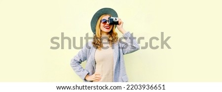 Portrait of stylish happy smiling woman with film camera taking picture wearing round hat, jacket coat on gray background