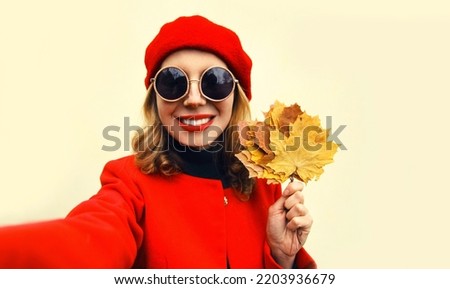 Autumn portrait of beautiful smiling woman stretching hand for taking selfie with smartphone with yellow maple leaves wearing red french beret on background