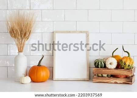 Picture frame mockup with wooden box of decorative pumpkins, vase of dry wheat on white table in nordic room interior.