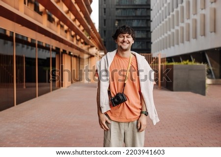Smiling young caucasian guy looks at camera standing against backdrop of city building. Brunette man with camera wears casual clothes. Happy weekend concept. Royalty-Free Stock Photo #2203941603