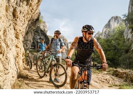 man caucasian male cyclist riding e-bike electric bicycle outdoor in mountain range at the gorge or ridge in front of his friends wear protective helmet and eyeglasses in sunny day copy space Royalty-Free Stock Photo #2203953717