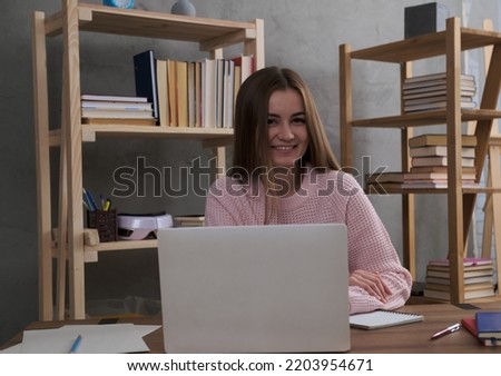 A cute young girl is working with a laptop at a desk. Many books on the shelves, in the background. Student time