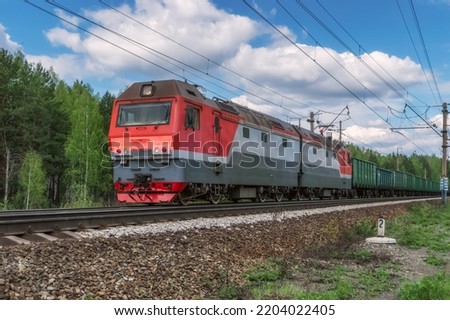A red electric locomotive with a freight train moves along the railroad tracks. Close-up. Freight train on the way next to the green forest against the blue sky. Russia, Ural
 Royalty-Free Stock Photo #2204022405