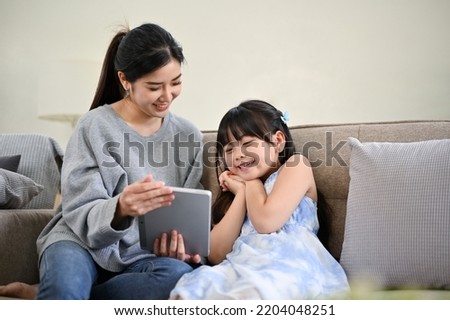A beautiful and kind Asian mom showing something on tablet to her daughter, watching kid's cartoon together, surprising her daughter with some picture on tablet screen. Happy family concept