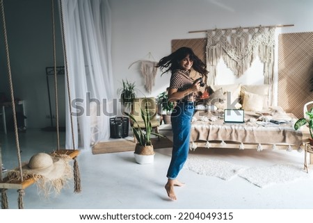 Side view of young female in casual clothes taking pictures with photo camera while jumping on floor near bed and potted plants in bedroom