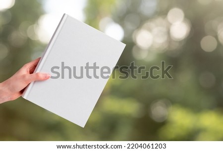 Hand holding book mockup on blurred background with green trees, grass. Literature about nature, ecology. Reading leisure, harmony concept. High quality photo