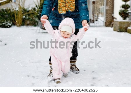 Adorable little baby girl making first steps outdoors in winter through snow. Cute toddler learning walking. Mother holding child on hand. Daughter and mum walk together. Happy family Royalty-Free Stock Photo #2204082625