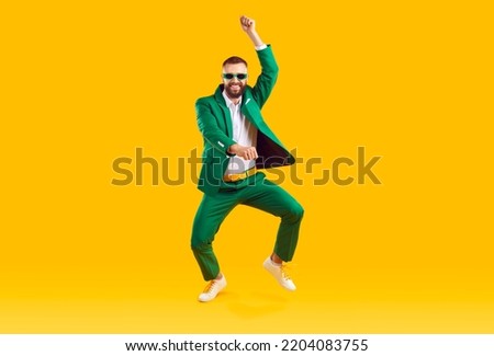 Cheerful energetic man has fun dancing in gangnam style at St. Patrick's Day party. Full length of man in trendy green suit and sunglasses of same color doing funny dance moves on orange background.