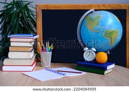 student home office table with white alarm clock, books, orange, colored notebooks, pencils in glass, chalk board, globe, white alarm clock, concept of education, back to school, knowledge day