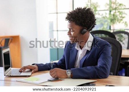 Portrait of smiling male office worker. Happy businessman with headphones signing documents, working on laptop. Portrait, office work concept.
